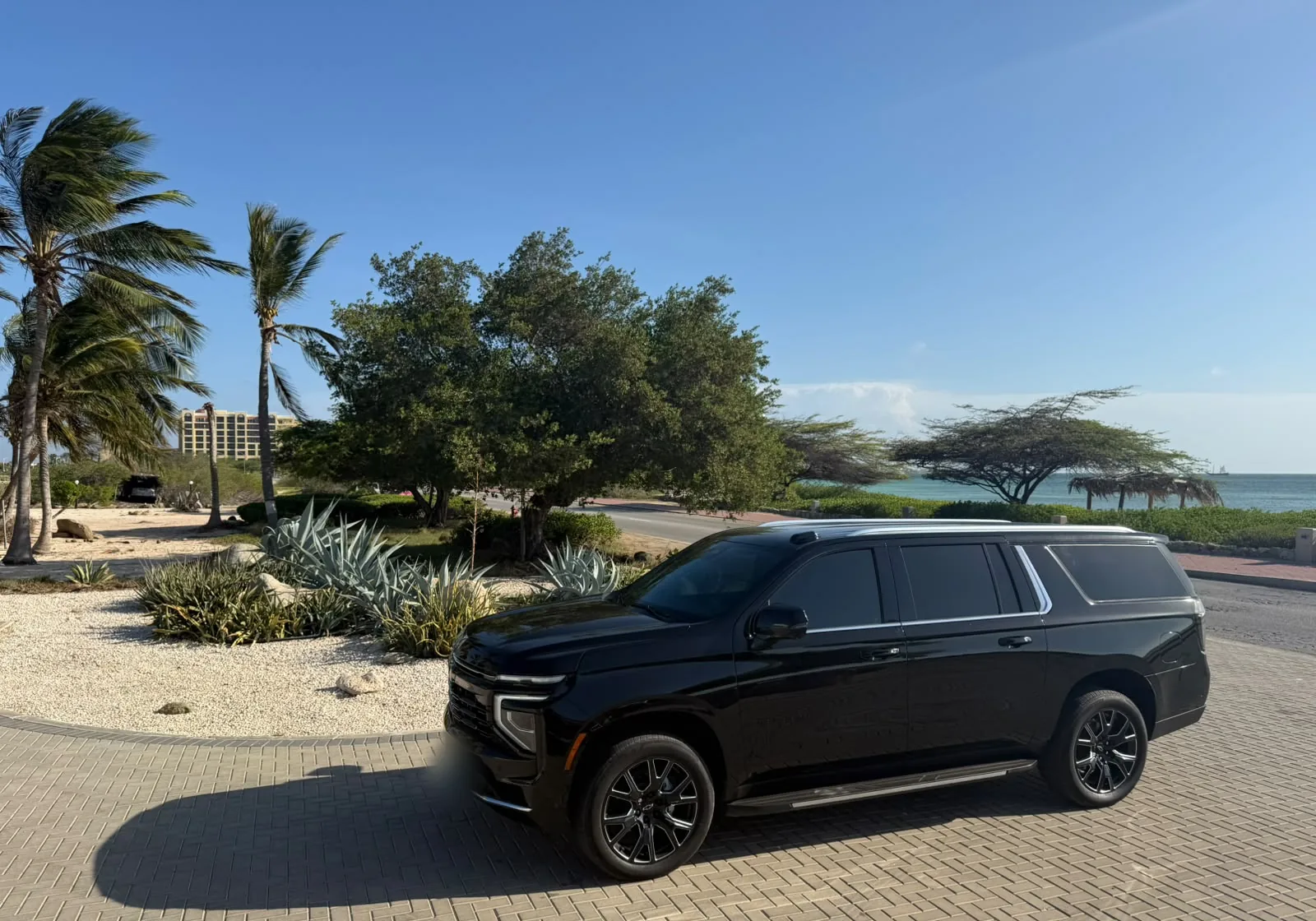 A black Chevrolet Suburban parked on a brick driveway beside desert plants and the sea in Aruba